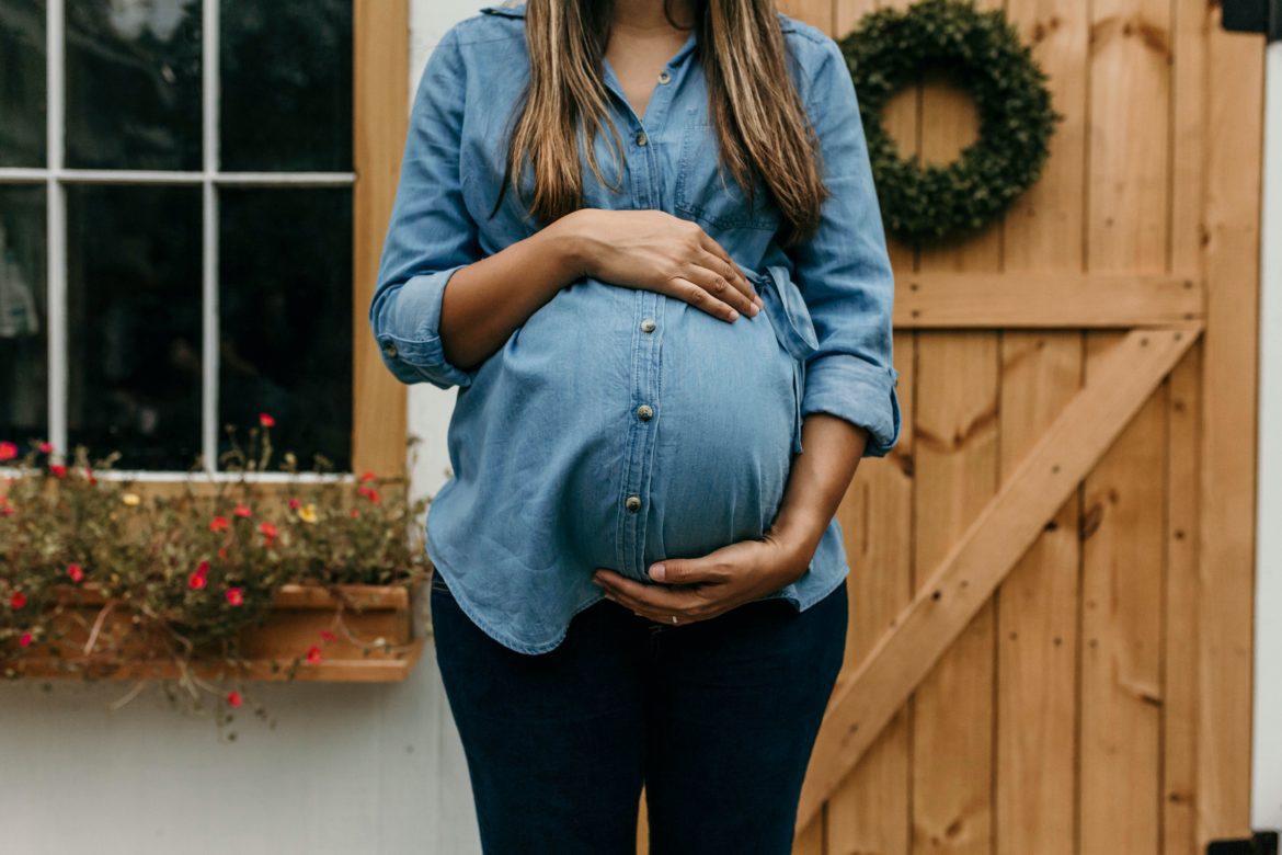 camylla-battani-dz8g0IxEqh0-unsplash Pregnant woman holding her stomach and standing in front of wooden house
